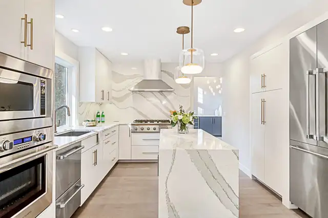 White kitchen with a waterfall quartz island, a full-height quartz backsplash, glass pendants and stainless wall ovens